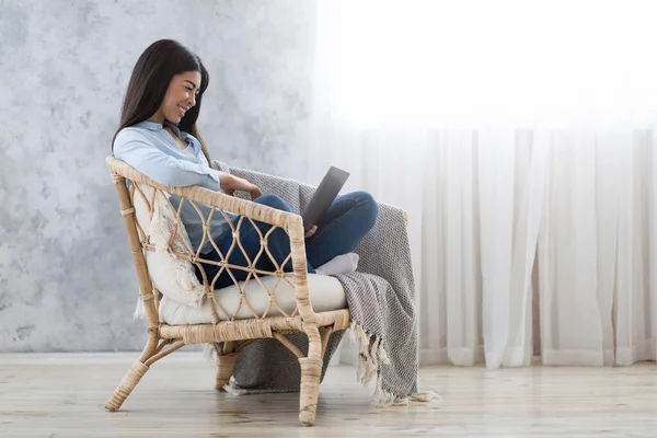 Lazer de fim de semana. Menina asiática relaxante com laptop na cadeira de vime em casa — Fotografia de Stock