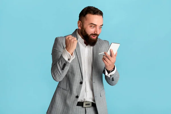 Opgewonden jonge zakenman op zoek naar smartphone en het maken van JA gebaar op blauwe achtergrond — Stockfoto