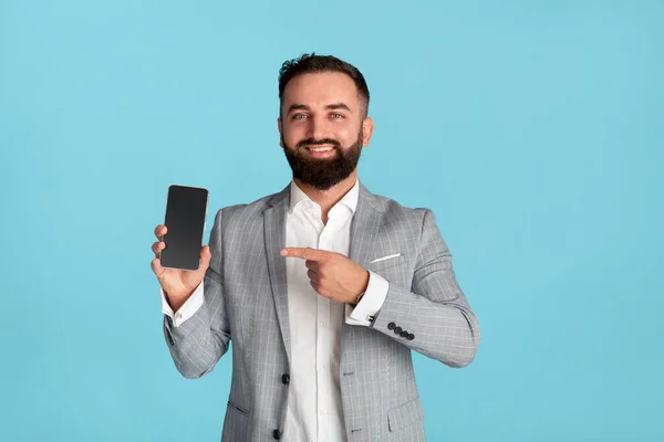 Glückliche Büroangestellte zeigt auf Handy mit leerem Bildschirm auf blauem Hintergrund, Platz für Design — Stockfoto