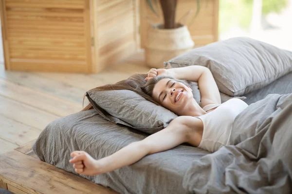 Mujer joven soñolienta despertando en su cama y estirándose en la mañana soleada — Foto de Stock