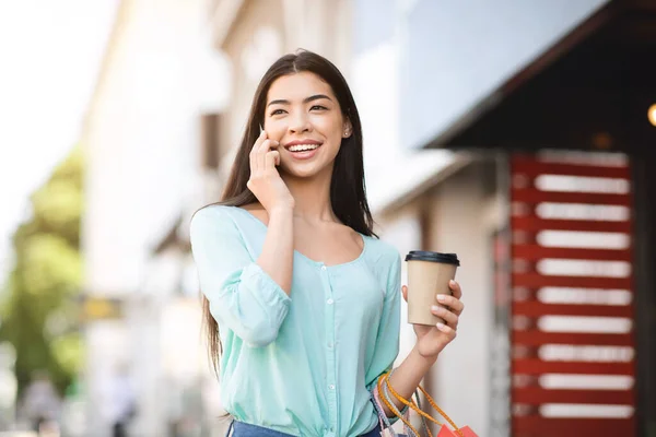 Joyful aziatische meisje met winkelen tassen en koffie praten op mobiele telefoon buiten — Stockfoto
