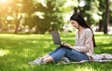 Gülümseyen Asyalı öğrenci laptopla dışarıda ders çalışıyor, sınavlara hazırlanıyor.