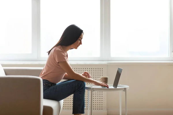 Libero professionista che lavora sul computer portatile seduto vicino alla finestra interna, vista laterale — Foto Stock