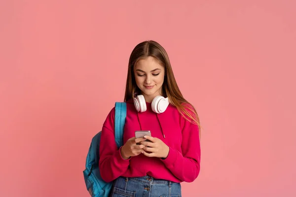 Moderne tieners. Glimlachend meisje met hoofdtelefoon en rugzak tikken op smartphone — Stockfoto