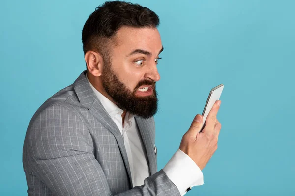 Workplace anger management. Pissed young businessman looking at smartphone screen on blue background — Stok Foto