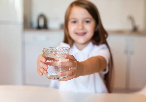 Menina mostrando vidro de água mineral bebendo sentado dentro de casa — Fotografia de Stock