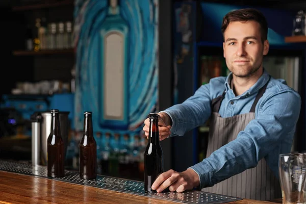 Serious handsome barman in apron opens bottle of beer in interior of modern pub — Stock Photo, Image