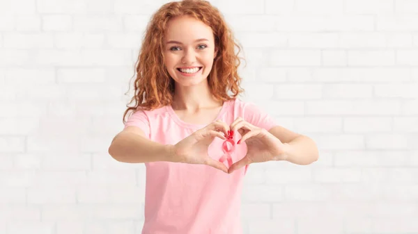 Girl Gesturing Heart Shape Framing Pink Ribbon Over White Wall