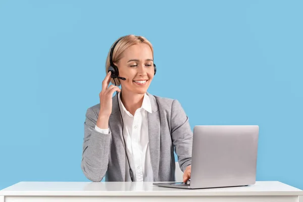 Mooie volwassen hulplijn operator met headset en laptop beantwoorden klanten bellen op de werkplek op blauwe achtergrond — Stockfoto