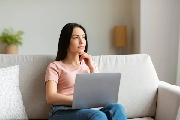 Pensive meisje op laptop denken zitten op bank thuis — Stockfoto