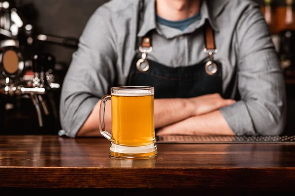 Barman em avental com braços cruzados no bar e atende cliente com copo de cerveja leve no interior do pub — Fotografia de Stock