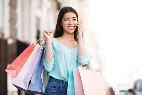 Llamando a Taxi. Joyful chica asiática sosteniendo bolsas de compras, Hablando en el teléfono celular al aire libre — Foto de Stock