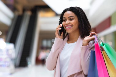 Alışveriş merkezinde elinde poşetlerle yürüyen ve cep telefonuyla konuşan zenci bir kadın.