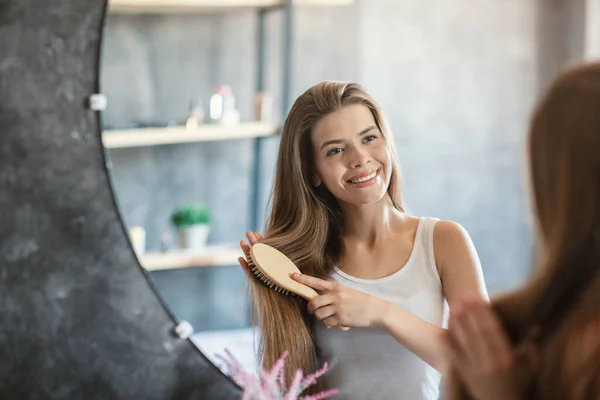 Lovely young girl brushing her beautiful long hair with wooden brush near mirror in bathroom — Stock Photo, Image