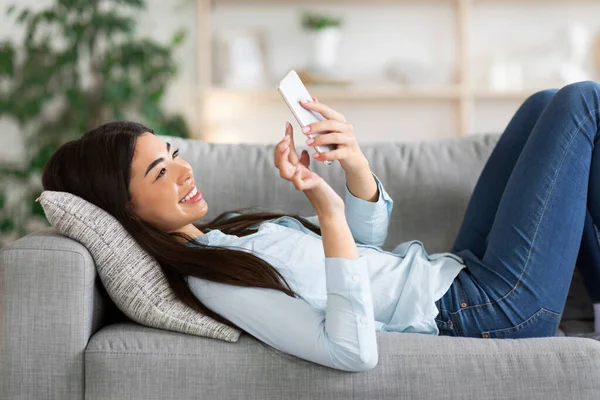 Asiática chica navegando por Internet en el teléfono inteligente mientras está acostado en el sofá en casa — Foto de Stock