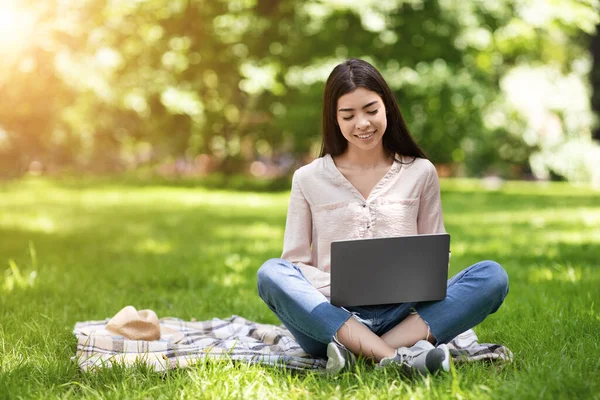 Freiberufliches Konzept. Lächelndes asiatisches Mädchen, das draußen am Laptop arbeitet und auf dem Rasen sitzt — Stockfoto