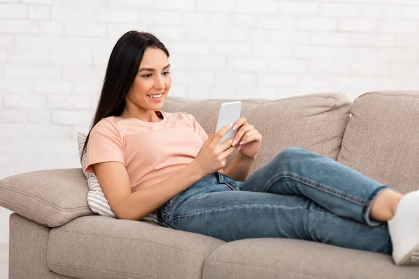 Mensajería de mujer sonriente en el teléfono móvil sentado en el sofá — Foto de Stock