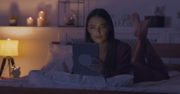 Серйозна жінка читає новини на цифровому планшеті, лежачи на ліжку — стокове відео