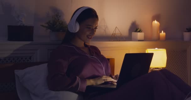 Молода дівчина в навушниках грає у відеоігри на ноутбуці в ліжку — стокове відео