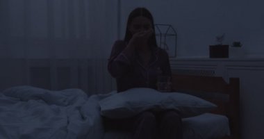 Genç bir kadın yatakta oturuyor, yatak odasında uyku hapları alıyor.