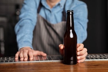 Önlüklü barmen ahşap tezgahta dinleniyor ve etiketsiz bir şişe bira servis ediyor.
