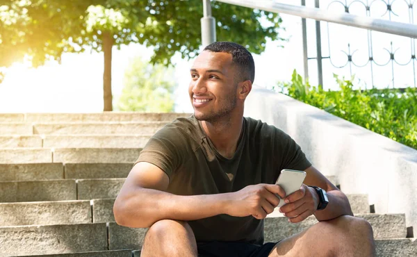 Glad afroamerikansk idrottsman använder smartphone på trappor under sin paus från träning i parken — Stockfoto