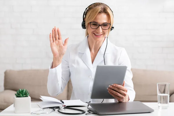 Videollamada al terapeuta. Mujer sonriente médico en auriculares saluda a la paciente y sostiene en la tableta —  Fotos de Stock