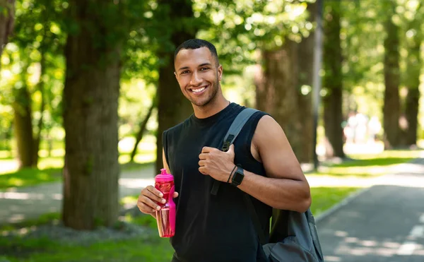 Retrato de deportista negro sonriente con mochila y botella de agua después de su entrenamiento en el parque —  Fotos de Stock