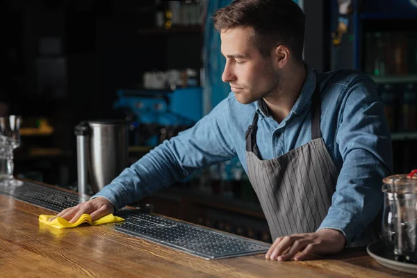 Kebersihan di pub. Bartender profesional berdiri di counter, menyeka permukaan kayu dengan kain — Stok Foto