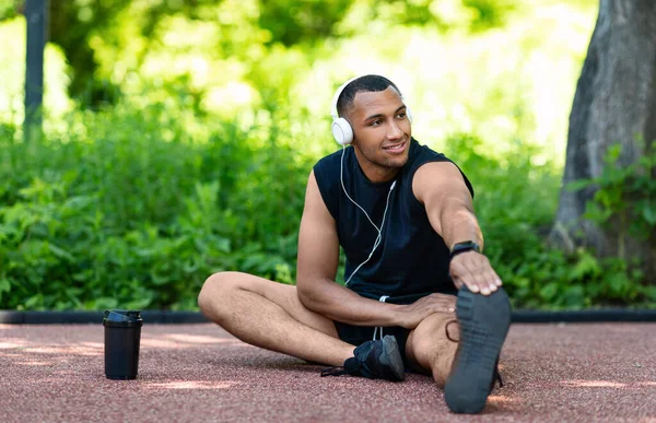 Attraktiver afroamerikanischer Typ, der Musik hört, während er sich nach seinem Lauf im Stadtpark dehnt, kopiert den Raum — Stockfoto
