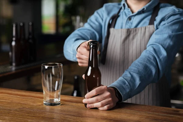 Szenische Kneipe und Brauerei. Barmann in Schürze öffnet Flasche Bier — Stockfoto