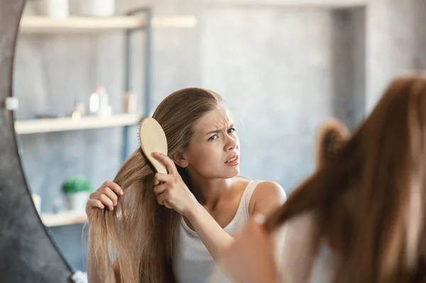 Frustrada jovem mulher tentando escovar seu cabelo emaranhado na frente do espelho no banheiro — Fotografia de Stock