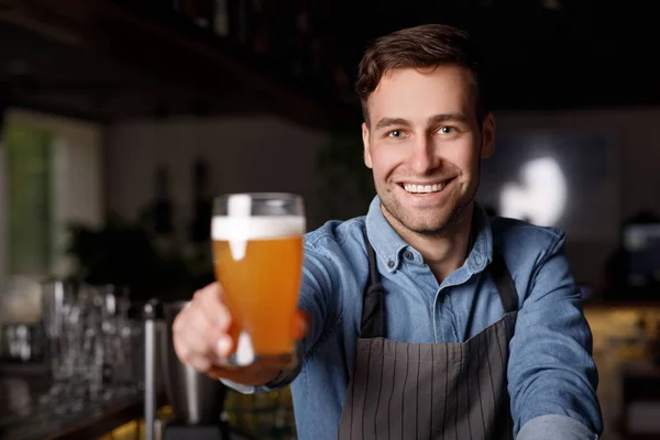 Trabajo en el pub. Sonriente barman guapo en delantal sostiene un vaso de cerveza — Foto de Stock