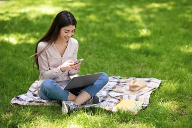 Genç Asyalı kadın akıllı telefondan mesaj atıyor ve dışarıda parkta dizüstü bilgisayar kullanıyor.