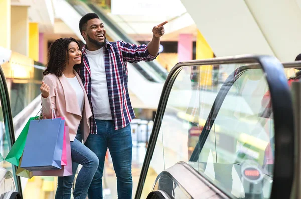 Negro millennial pareja subiendo la escalera mecánica en el centro comercial y señalando a un lado — Foto de Stock
