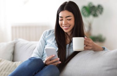 Ev eğlencesi. Gülümseyen Asyalı kız akıllı telefon ve kahveyle kanepede dinleniyor.