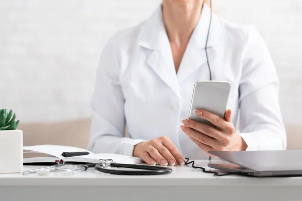 Επίσκεψη στο γιατρό. Γιατρός κοιτάζοντας smartphone στην επιφάνεια εργασίας, κοντά είναι στηθοσκόπιο σημειωματάριο και φορητό υπολογιστή — Φωτογραφία Αρχείου