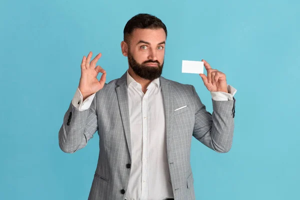 Empleado corporativo de confianza sosteniendo la tarjeta de visita vacía y mostrando un gesto aceptable sobre fondo azul, espacio para el diseño — Foto de Stock
