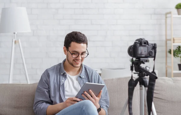 Revisión en video blog. Chico en gafas sostiene la tableta digital en las manos, se sienta en el sofá con cámara de vídeo — Foto de Stock