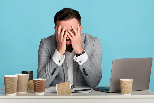 Vyčerpaný podnikatel se snaží nespát na pracovišti s prázdnými šálky kávy a notebook na modrém pozadí — Stock fotografie