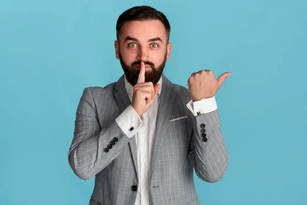 Attraente imprenditore mostrando gesto HUSH e indicando da parte su sfondo blu — Foto Stock