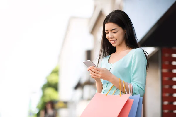 Moda satışları ve anlaşmaları. Alışveriş çantalı Asyalı kız dışarıda akıllı telefon kullanıyor. — Stok fotoğraf