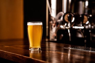 Bardakta hafif bira. Kahverengi ahşap tezgahta köpüklü bira, yanında metal musluklar.