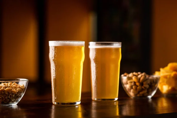 Ahşap masada iki bardak bira, yanında fıstık, fındık ve cips var. — Stok fotoğraf