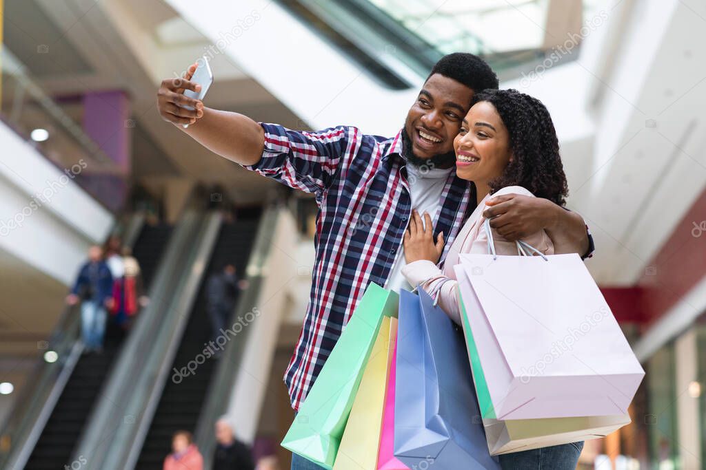 Shopping Fun. Joyful black couple taking selfie in mall, holding shopper bags