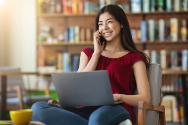 Laptopta çalışan ve kafede cep telefonuyla konuşan gülümseyen Asyalı kadın.