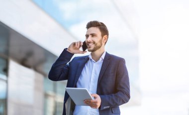 Meşgul genç girişimci cep telefonuyla iletişim kuruyor ve şehirdeki tablet bilgisayardaki belgelere bakıyor.