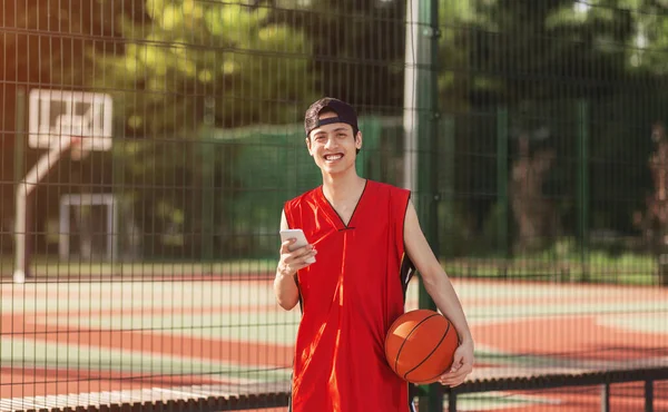 Sorrindo asiático basquetebolista com telefone celular posando na arena ao ar livre após o jogo — Fotografia de Stock