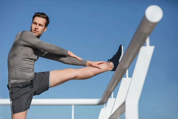 Millennial guy in sportswear doing stretching, on blue sky background