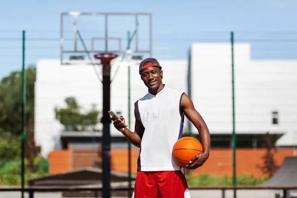 Happy black basketballer dengan ponsel berdiri di arena permainan luar ruangan, ruang kosong — Stok Foto